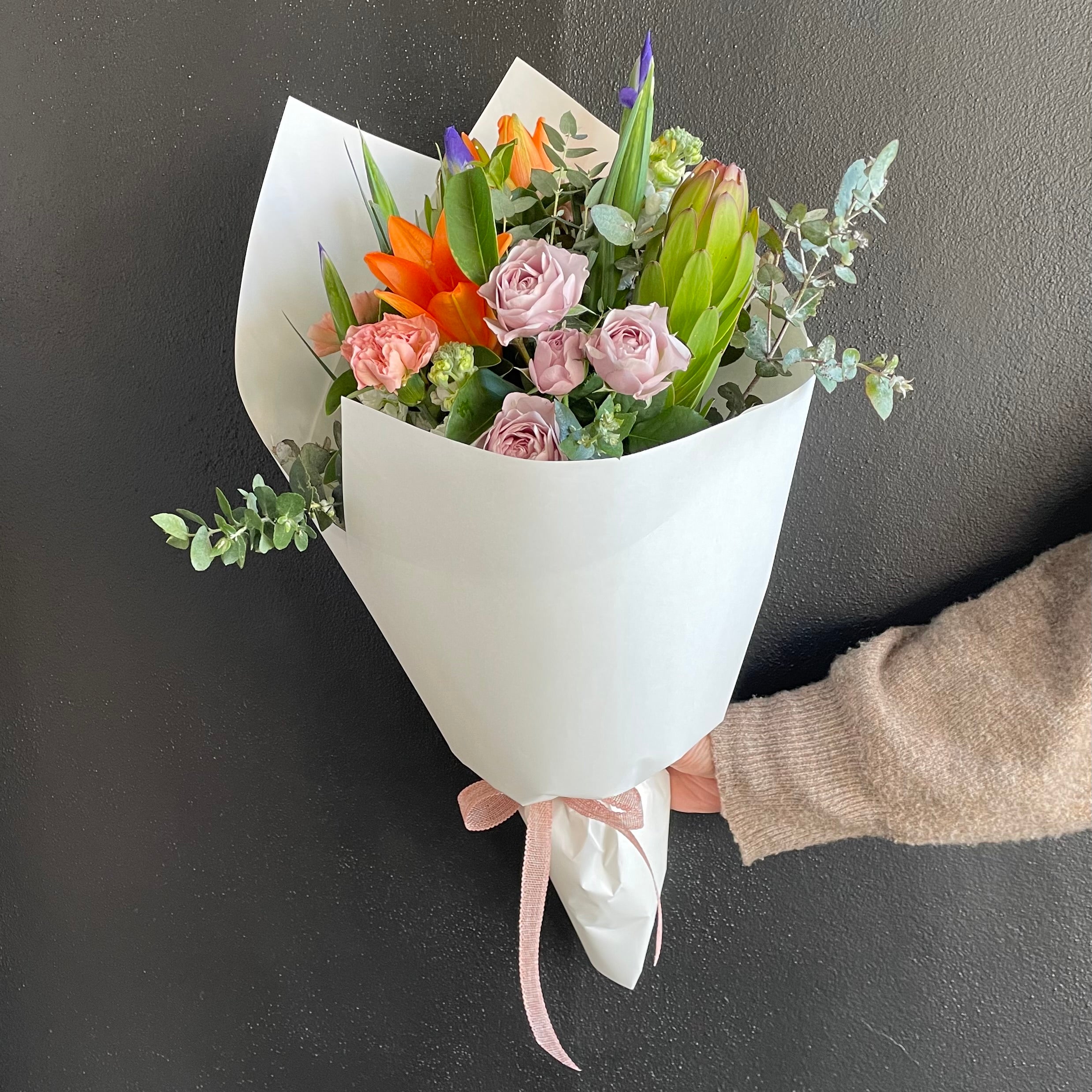 Flower bouquet with bright coloured orange, purple and pink flowers wrapped in white Kraft paper with pink bow. Bouquet designed by Bendigo Florist Blumetown. 